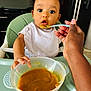baby, high_chair, baby_food, spoon, hand, feeding, bib, curly_hair, indoor, table, container, person, child, mealtime, cute, expression, food, kitchen, nurturing, feeding_time