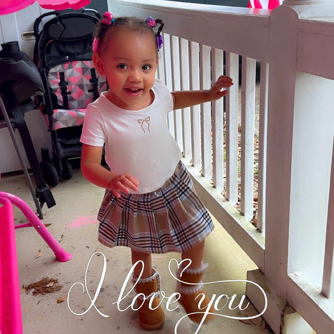 Julianna is registered to the contest to win money with this photo: toddler, child, porch, smiling, plaid_skirt, boots, white_tshirt, hair_buns, stroller, car, railing, outdoor, concrete_floor, pink, love_text, happy, cute, playful, daylight, person