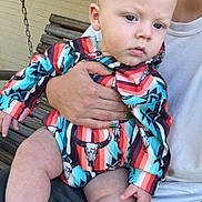 Rylan is registered to the contest to win money with this photo: baby, child, person, holding, colorful_clothing, striped_outfit, bull_skull_pattern, hand, nail_polish, green_nails, wooden_bench, swing, outdoor, brick_wall, curious_expression, blond_hair, mohawk, skin, legs, casual