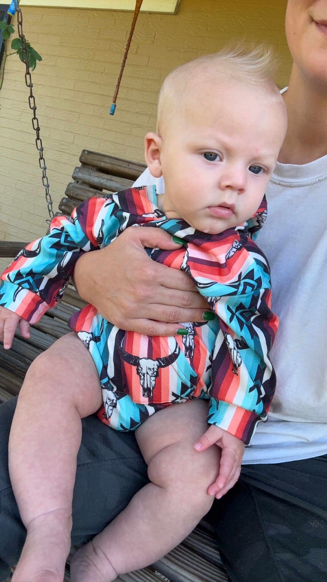 Rylan is registered to the contest to win money with this photo: baby, child, person, holding, colorful_clothing, striped_outfit, bull_skull_pattern, hand, nail_polish, green_nails, wooden_bench, swing, outdoor, brick_wall, curious_expression, blond_hair, mohawk, skin, legs, casual