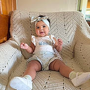 Astrid is registered to the contest to win money with this photo: baby, child, smiling, headband, clothing, knitted_blanket, chair, indoor, cute, happy, infant, person, footwear, socks, white_shoes, wall, light_switch, wood_panel, seated, portrait