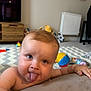baby, tongue, playful, indoor, toys, living_room, child, furniture, floor, carpet, radiator, colorful, expression, cute, person, hand, face, skin, hair, fun