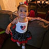 Ariana joined the competition — help win amazing prizes! child, toddler, girl, smiling, portrait, indoor, living_room, couch, furniture, dress, skirt, hair_bows, patriotic, american_flag, red_white_blue, socks, standing, home, dining_area, baby_seat