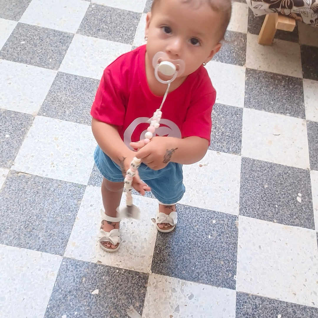 Lilia participe au concours pour gagner de l'argent avec cette photo : toddler, child, pacifier, pigtails, red_shirt, denim_shorts, sandals, floor, checkered_floor, indoor, curious, standing, looking_up, beaded_clip, furniture, sofa, pattern, young_child, cute, portrait