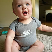 Braxton is registered to the contest to win money with this photo: baby, child, infant, onesie, grey_clothing, nike_logo, blue_eyes, sitting, blanket, indoor, wooden_floor, expression, cute, happy, portrait, skin, person, head, hand, foot