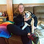 black_clothing, blanket, brown_couch, casual_clothing, comfortable, couch, dog, footwear, happy_dog, home, indoor, leisure, pet, rainbow_blanket, relaxed, seated, shoe, smiling, woman, wood_paneling