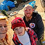 child, adult, family, outdoor, beanie, plaid_shirt, sippy_cup, smiling, beard, blonde_hair, piercing, hoodie, casual_clothing, backyard, garden_tools, sunlight, dirt_ground, container, plastic_bin, selfie
