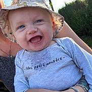 Jaïden a rejoint le concours — aidez-le/la à gagner de superbes lots ! baby, child, smiling, blue_eyes, hat, bucket_hat, shirt, text_on_clothing, outdoor, sunlight, person, happy, holding, nature, greenery, arm, bracelet, portrait, cute, infant