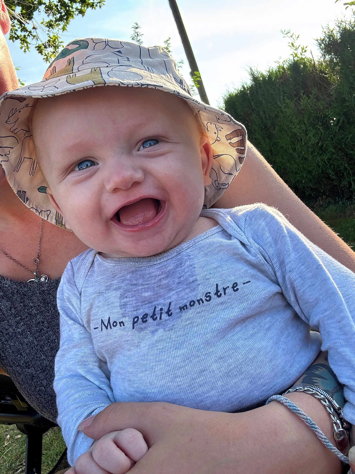 Jaïden a rejoint le concours — aidez-le/la à gagner de superbes lots ! baby, child, smiling, blue_eyes, hat, bucket_hat, shirt, text_on_clothing, outdoor, sunlight, person, happy, holding, nature, greenery, arm, bracelet, portrait, cute, infant