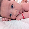baby, infant, face, blue_eyes, hand, skin, red_bracelet, lying_down, white_quilt, close_up, portrait, child, cute, resting, person, indoor, soft_light, curly_hair, expression, human