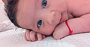Maria-Cezara joined the competition — help win amazing prizes! baby, close_up, blue_eyes, curly_hair, red_bracelet, hand, face, skin, lying_down, white_quilt, infant, cute, portrait, child, resting, soft_light, indoors, human, expression, newborn