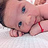 baby, close_up, blue_eyes, curly_hair, red_bracelet, hand, face, skin, lying_down, white_quilt, infant, cute, portrait, child, resting, soft_light, indoors, human, expression, newborn