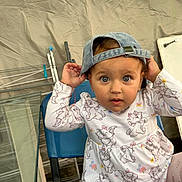 Stefania is registered to the contest to win money with this photo: toddler, child, cap, denim_cap, blue_chair, cartoon_clothing, long_sleeve_shirt, curious_expression, indoor, face, person, seated, patterned_clothing, cute, baby, hands_up, light_brown_hair, large_eyes, playful, casual