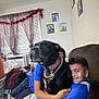 air_conditioner, black_dog, boy, casual_clothing, chain_collar, child, couch, curtains, dog, family_photos, furniture, garland, hugging, indoor, laundry_basket, living_room, messy_room, smiling, thumb, window
