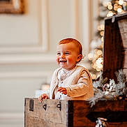 Lizio participe au concours pour gagner de l'argent avec cette photo : toddler, child, smiling, wooden_chest, holiday_decor, candles, pine_tree, indoor, warm_lighting, rosy_cheeks, vest, long_sleeve_shirt, decorative_plants, blurred_background, festive, cute, portrait, cozy, happy, holiday_season