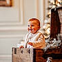 toddler, child, smiling, wooden_chest, holiday_decor, candles, pine_tree, indoor, warm_lighting, rosy_cheeks, vest, long_sleeve_shirt, decorative_plants, blurred_background, festive, cute, portrait, cozy, happy, holiday_season