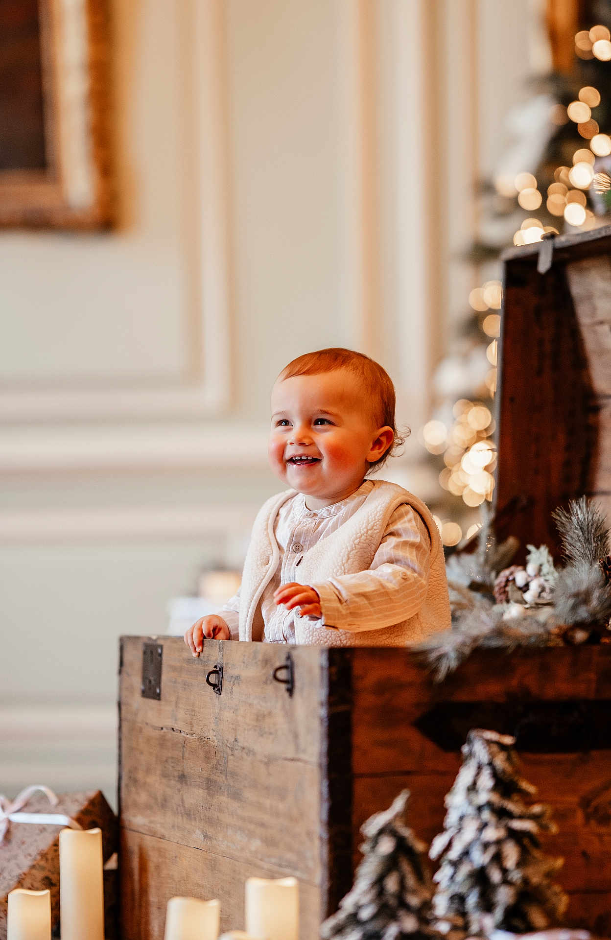 Lizio participe au concours pour gagner de l'argent avec cette photo : toddler, child, smiling, wooden_chest, holiday_decor, candles, pine_tree, indoor, warm_lighting, rosy_cheeks, vest, long_sleeve_shirt, decorative_plants, blurred_background, festive, cute, portrait, cozy, happy, holiday_season
