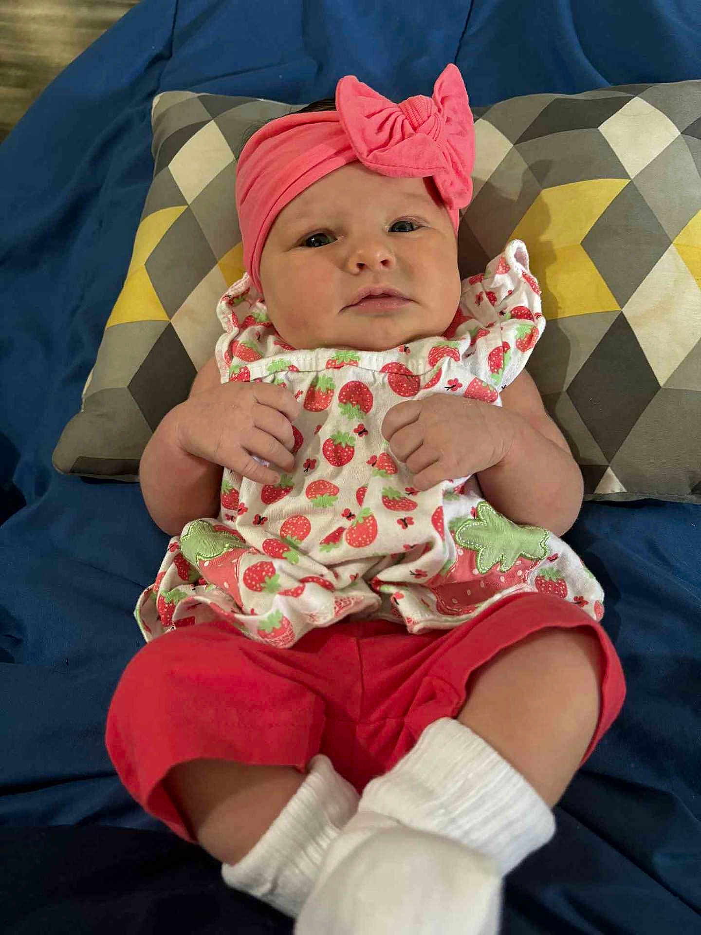 Alexis is registered to the contest to win money with this photo: baby, infant, headband, pink, bow, strawberry_pattern, clothing, pillow, blanket, red_pants, white_socks, cute, portrait, indoor, child, person, lying_down, soft_fabric, colorful, comfortable