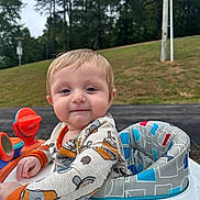 Darren joined the competition — help win amazing prizes! baby, child, infant, portrait, smiling, outdoor, activity_seat, toy, playground, grass, trees, park, road, lamppost, clothing, onesie, orange, blue, sitting, face