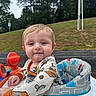baby, child, infant, portrait, smiling, outdoor, activity_seat, toy, playground, grass, trees, park, road, lamppost, clothing, onesie, orange, blue, sitting, face