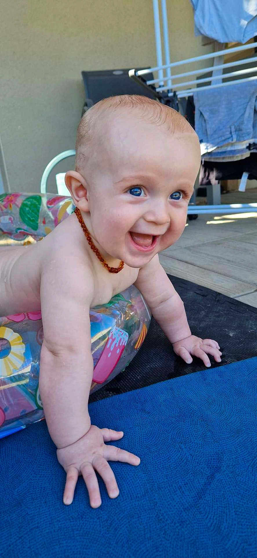 Lewenn participe au concours pour gagner de l'argent avec cette photo : arm, baby, background, blue_eyes, child, clothing, floor, hand, happy, head, infant, inflatable_ring, laundry, necklace, outdoor, person, playful, skin, smiling, sunlight