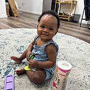 Denver is registered to the contest to win money with this photo: baby, child, smiling, sitting, toy, rug, floor, snack_container, purple_phone_case, indoor, wooden_floor, furniture, home, cute, person, earrings, clothing, happy, portrait, play