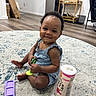baby, child, smiling, sitting, toy, rug, floor, snack_container, purple_phone_case, indoor, wooden_floor, furniture, home, cute, person, earrings, clothing, happy, portrait, play