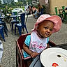 Denver is registered to the contest to win money with this photo: toddler, child, pink_hat, bib, high_chair, outdoor, cafe, table, plate, strawberry, curious_expression, urban, people, chairs, concrete, daylight, casual, person, food, smile