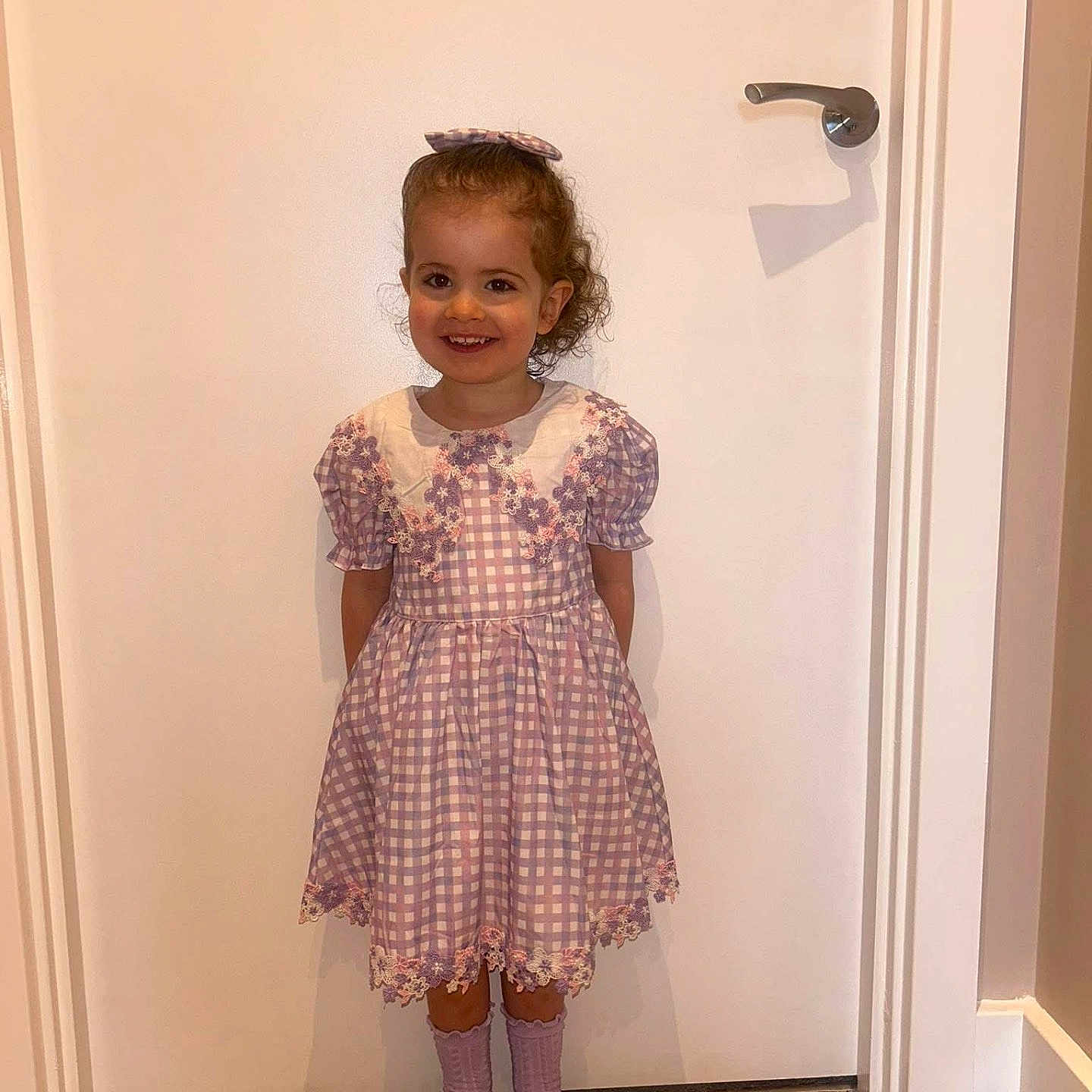 Sydney is registered to the contest to win money with this photo: bow, checkered, child, curly_hair, cute, door, dress, fashion, girl, happy, indoors, portrait, purple, shoes, smiling, socks, standing, white_door, wooden_floor, young