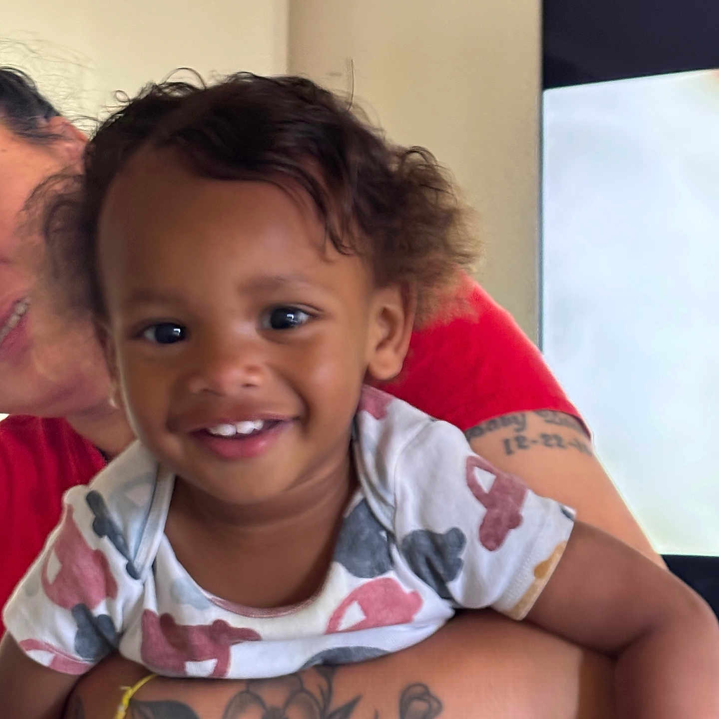 Carnello is registered to the contest to win money with this photo: adult, arm, child, closeup, curly_hair, face, happy, holding, human, indoor, patterned_shirt, person, portrait, red_clothing, skin, smiling, tattoo, television, toddler, white_shirt