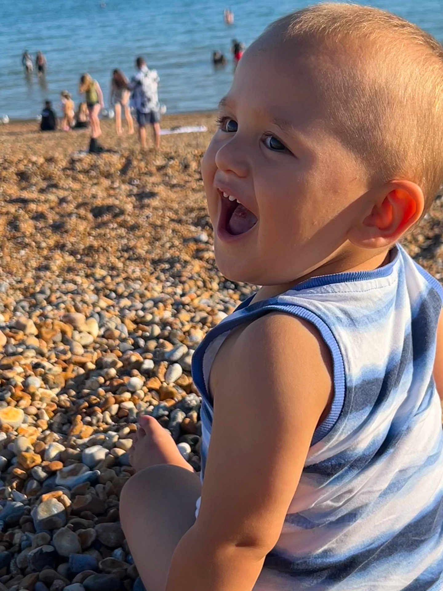 Tom is registered to the contest to win money with this photo: toddler, child, smiling, beach, pebbles, summer, sea, water, sunlight, casual_clothing, outdoor, happy, person, nature, vacation, relaxation, fun, family, daytime, shore