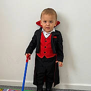 Tom is registered to the contest to win money with this photo: black_jacket, boots, carpet, child, clothing, colorful_mat, costume, floor, indoor, person, playful, portrait, red_vest, serious_expression, short_hair, standing, toddler, toy_cane, vampire, wall