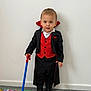 black_jacket, boots, carpet, child, clothing, colorful_mat, costume, floor, indoor, person, playful, portrait, red_vest, serious_expression, short_hair, standing, toddler, toy_cane, vampire, wall