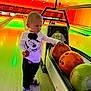 bowling_alley, bowling_balls, casual_clothing, child, colorful, curious, entertainment, floor, indoor, lanes, mickey_mouse, neon_lights, person, recreation, sandals, shirt, sport, standing, toddler, toy