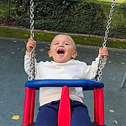 Tom joined the competition — help win amazing prizes! child, toddler, swing, playground, outdoor, park, happy, smiling, clothing, white_sweater, navy_pants, chains, red_swing, blue_swing, trees, hedge, people, grass, daylight, fun