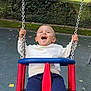blue_swing, chains, child, clothing, daylight, fun, grass, happy, hedge, navy_pants, outdoor, park, people, playground, red_swing, smiling, swing, toddler, trees, white_sweater