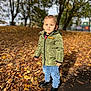 autumn, background, blue_jeans, boots, casual_clothing, child, daylight, fall, green_jacket, leaf_litter, leaves, nature, outdoor, park, person, seasonal, standing, sunlight, trees, young_child