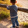 Adélio participe au concours pour gagner de l'argent avec cette photo : child, toddler, back_view, striped_sweater, jeans, white_sneakers, wooden_fence, beach, sand, grass, sunlight, shadow, outdoor, nature, water, sky, summer, casual_clothing, young_child, standing
