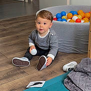 Adélio participe au concours pour gagner de l'argent avec cette photo : toddler, child, indoor, wooden_floor, ball, playpen, colorful_balls, shoe, rug, blanket, card, striped_shirt, black_pants, curious, sitting, playtime, home, person, floor, casual_clothing