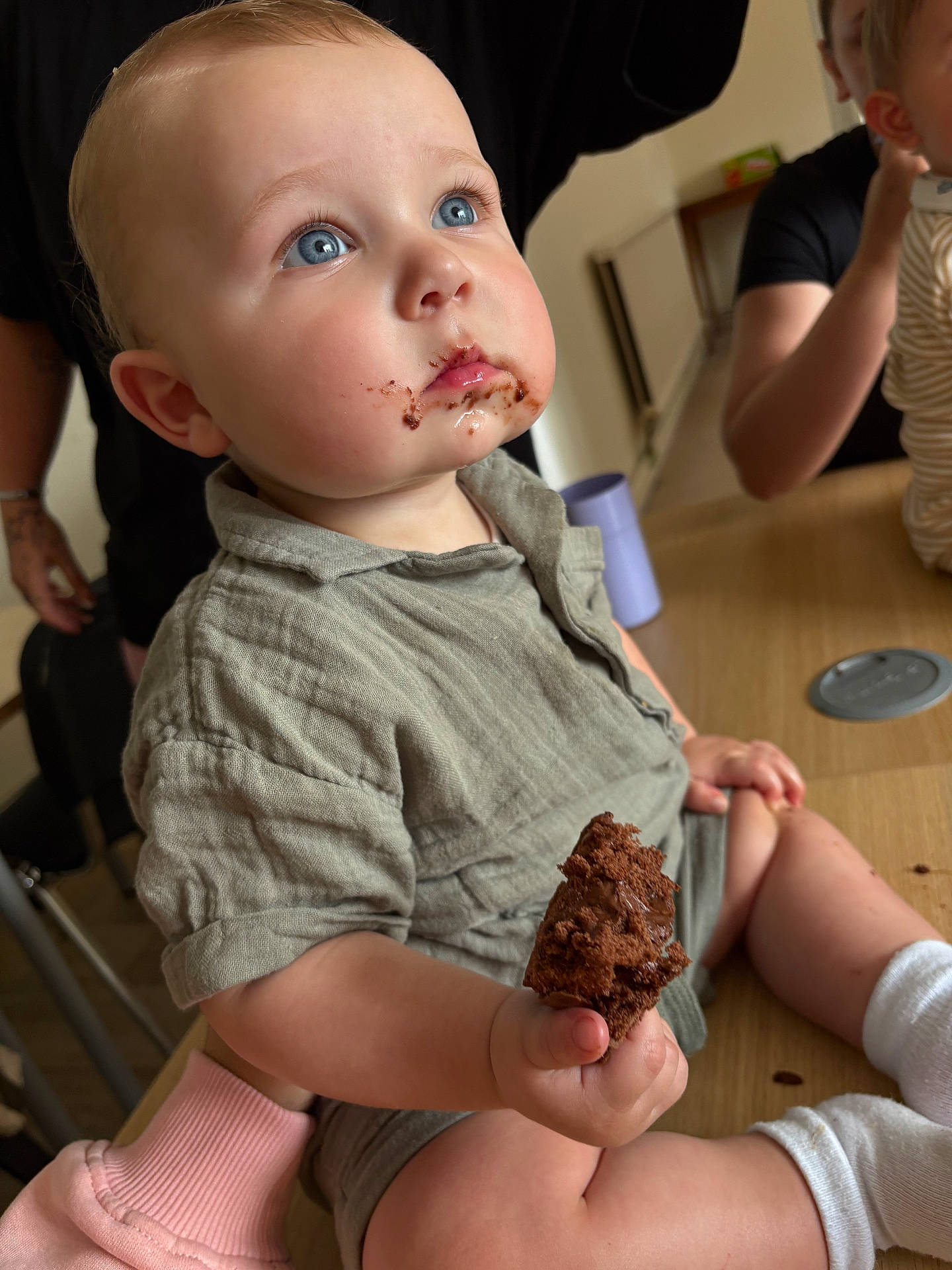 adult, baby, blue_eyes, child, chocolate, clothing, curious, cute, face, food, furniture, hand, indoor, mouth, muffin, person, sitting, socks, supporting_hand, table