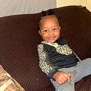 Armani is registered to the contest to win money with this photo: child, toddler, smiling, sitting, couch, brown_couch, leopard_print, black_clothing, gray_pants, happy, indoors, hairstyle, cute, person, portrait, furniture, young_child, bright, casual_wear, cozy