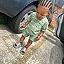 toddler, child, cornrows, green_shirt, shorts, parking_lot, car, tire, sidewalk, shoes, smile, outdoor, daylight, person, young_child, casual_clothing, earrings, concrete, vehicle, standing