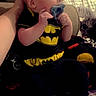 baby, background, batman, black_clothing, blanket, candid, child, comfort, cute, fan, footwear, hand, head, indoor, infant, pacifier, person, sitting, socks, support
