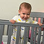 baby, crib, onesie, baby_clothes, hand, railing, pink_blanket, indoor, child, smiling, face, person, bedroom, furniture, happy, standing, infant, home, cute, playful