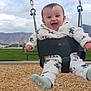 baby, child, swing, playground, outdoor, smiling, happy, clothing, jacket, mountains, sky, clouds, woodchips, seat, chains, bench, grass, person, feet, sock