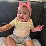 baby, child, smile, headband, pink_bow, shirt, yellow_shirt, text_on_clothing, couch, blanket, indoor, happy, cute, person, infant, seated, furniture, soft_texture, chubby_cheeks, casual_clothing