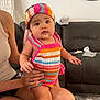 baby, child, infant, colorful_clothing, headband, knitted_outfit, person, sitting, indoor, couch, adult_hand, living_room, mirror, towel, curious_expression, skin, arm, leg, furniture, casual
