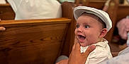 Myles joined the competition — help win amazing prizes! baby, child, infant, smiling, happy, person, hat, white_clothing, indoor, church, wooden_pews, adult_hand, portrait, seated, people, family, celebration, cute, bright_eyes, holding