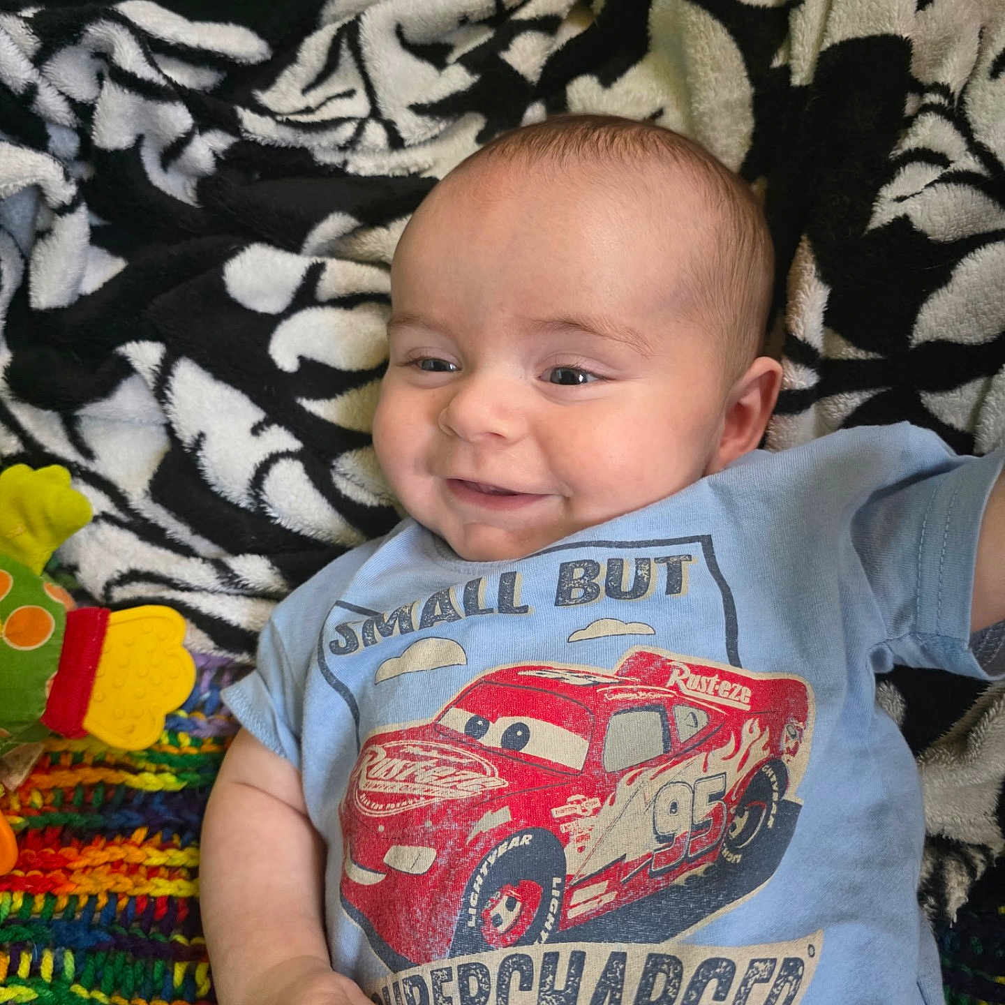 Scotty is registered to the contest to win money with this photo: animated_character, baby, blanket, blue_tshirt, child, clothing, colorful, cute, face, happy, indoor, infant, knitted_blanket, lying_down, patterned_blanket, person, playful, racecar, smiling, toy