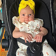 Olympe participe au concours pour gagner de l'argent avec cette photo : baby, child, infant, stroller, yellow_bow, headband, white_outfit, pineapple_pattern, black_stroller, seatbelt, hand, face, eyes, cute, indoors, person, portrait, sitting, clothing, soft_lighting